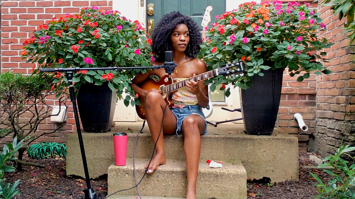 Baltimore, Maryland musician Evan Nicole Bell playing guitar outside between two flower bushes. She is playing an Epiphone Les Paul, and behind her is a Fender Jazz Bass.